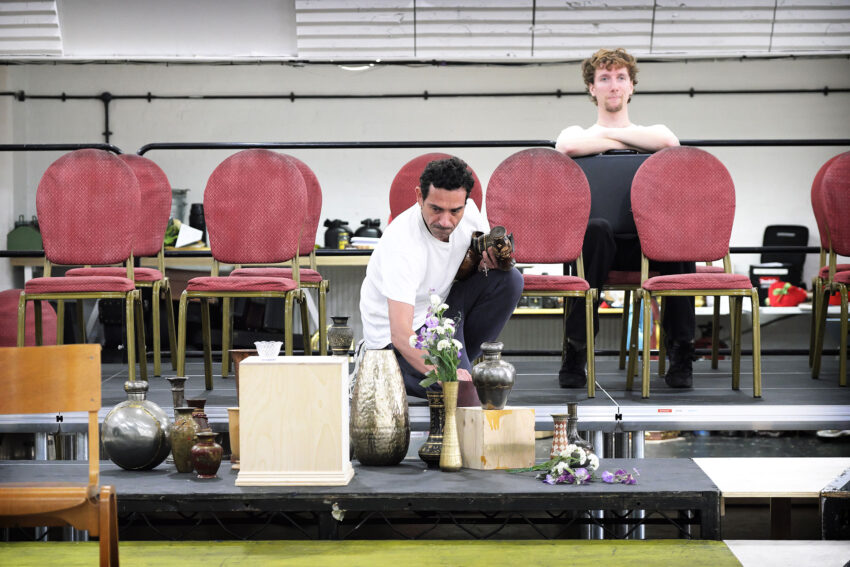 A man arranges vases and flowers on a stage while another person sits behind stacked red chairs, leaning forward and watching. The setting appears to be a rehearsal or stage set-up.
