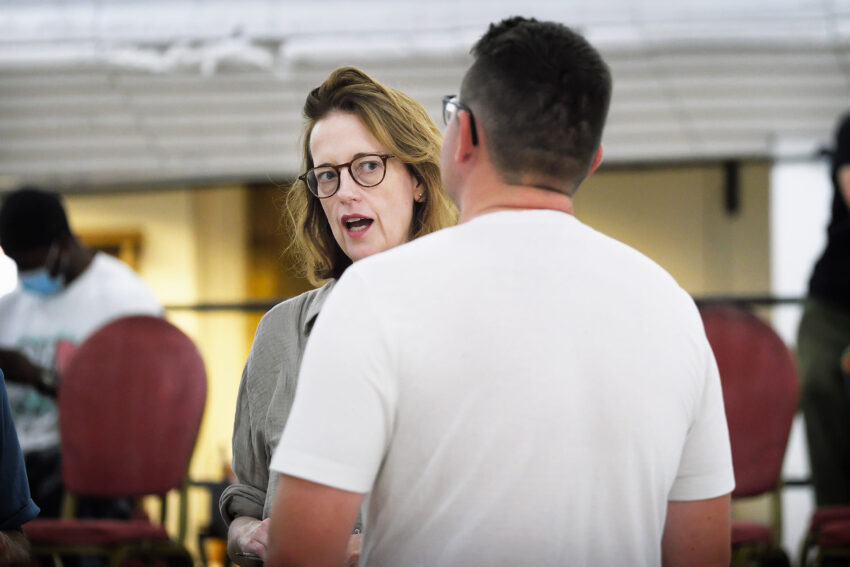 A woman wearing glasses speaks to a man in a white shirt. Both are indoors, with red chairs and other people blurred in the background.