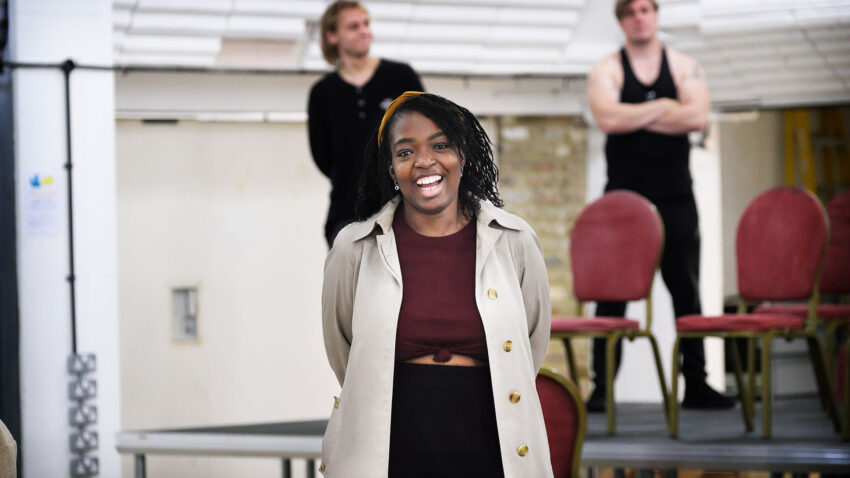 A woman smiles whilst standing in front of two people and some stacked chairs on a platform in a brightly lit indoor space.