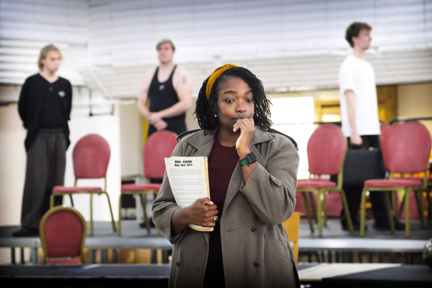 A woman in a coat holds a script and looks thoughtful. Three people stand behind her on a stage with red chairs, some wearing casual clothes. The setting suggests a theatrical rehearsal.