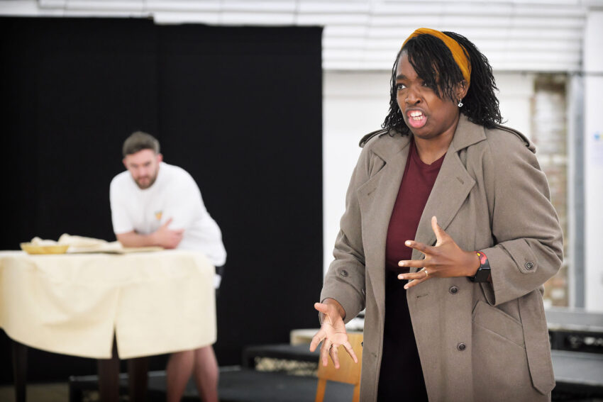 A woman in a brown coat gestures passionately whilst speaking. In the background, a man in white leans on a table covered with a cream cloth, watching her. The scene appears to take place indoors, during a rehearsal.