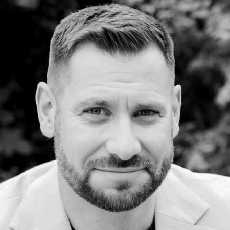 Black and white portrait of a man with short hair and a beard, wearing a light-coloured jacket, looking slightly up at the camera with a soft smile. The background is blurred with foliage.