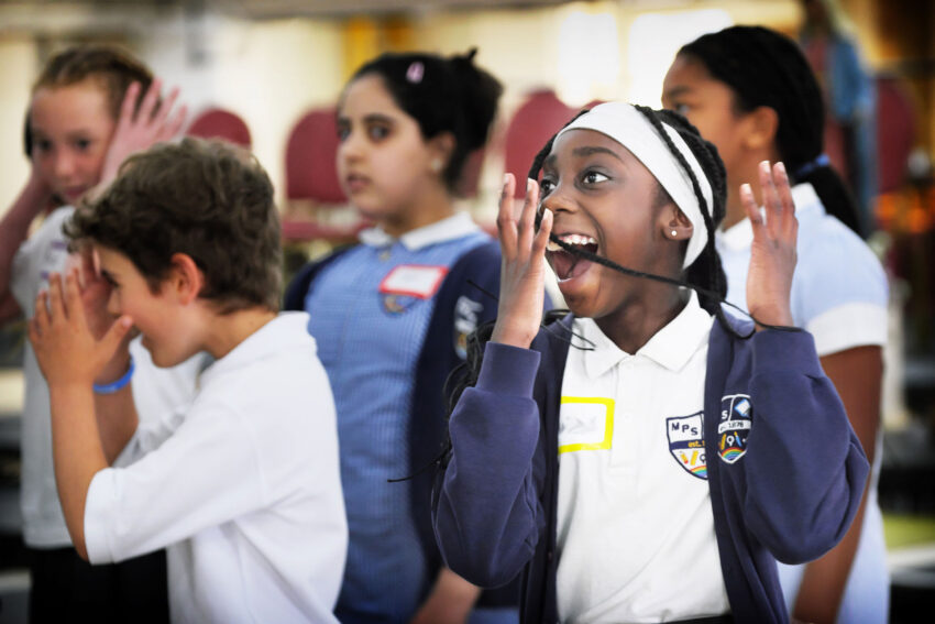 A group of children in school uniforms stand together indoors, some with hands on their faces, expressing excitement or surprise. One girl in the foreground smiles widely with her hands by her cheeks.