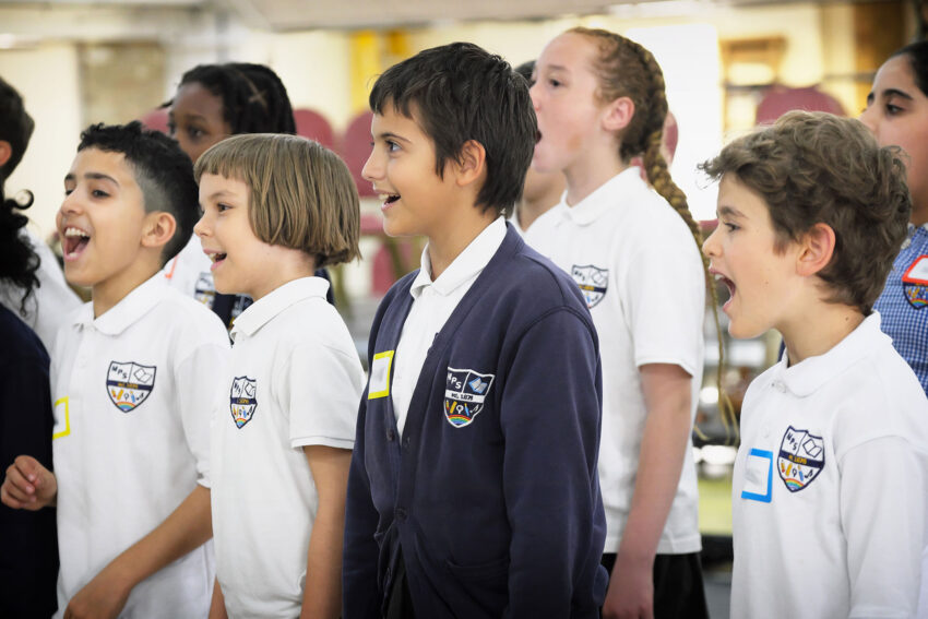 A group of school children wearing uniforms stand in a row, smiling and singing enthusiastically in a rehearsal setting.