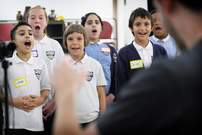 A group of children in school uniforms sing together whilst a conductor, seen from behind, leads them. The children appear enthusiastic and focused, with name tags on their shirts.