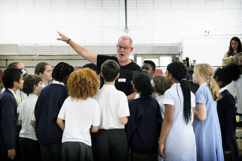 A man stands in the centre of a group of school children, gesturing and speaking animatedly as they listen attentively in a brightly lit classroom.