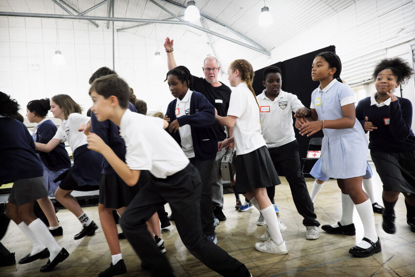 A group of schoolchildren and an adult move energetically indoors, some smiling and laughing, in a bright room with high ceilings and visible lighting. Everyone appears to be participating in an activity together.
