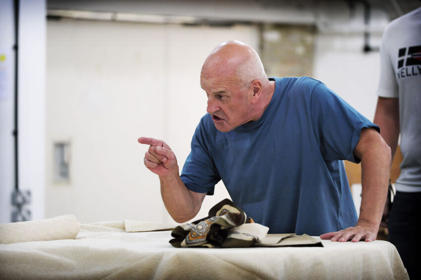 A bald man in a blue shirt points and speaks emphatically whilst standing by a table covered with fabric, with another person partially visible beside him in a workshop setting.
