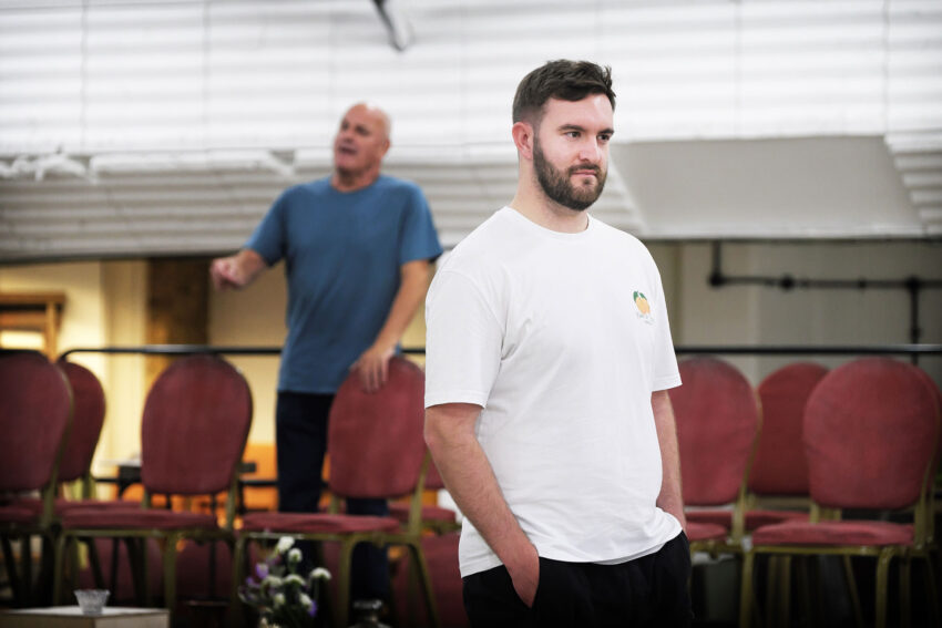 A man in a white T-shirt stands with his hands in his pockets, looking ahead. Behind him, another man in a blue shirt appears to be speaking or singing. Rows of red chairs are stacked in the background.