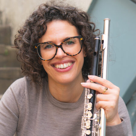 A woman with curly hair and glasses smiles while holding a flute. She is wearing a grey top and is sitting on stairs, with a light blue handrail in the background.