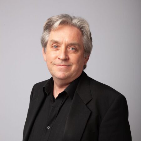 A man with grey hair wearing a black dress shirt and black blazer stands in front of a light grey background, smiling softly at the camera.
