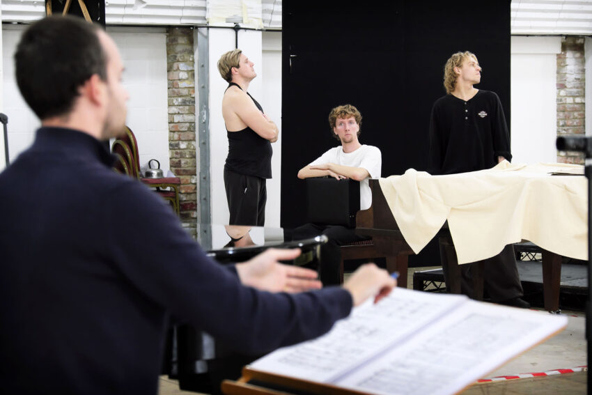Four people rehearse in a studio. One person conducts in the foreground, while three others stand or sit near a piano covered with a white cloth, appearing thoughtful and attentive. Brick walls and music sheets are visible.