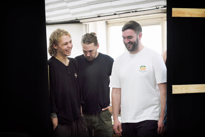Three men stand together indoors, smiling and laughing. Two wear dark shirts, one wears a white shirt with a small orange fruit logo. Bright daylight comes through windows behind them. The mood appears light and friendly.