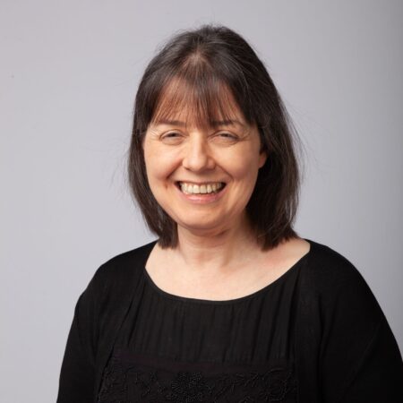 A woman with shoulder-length brown hair and a fringe smiles at the camera. She is wearing a black top with a cardigan. The background is plain and light grey.
