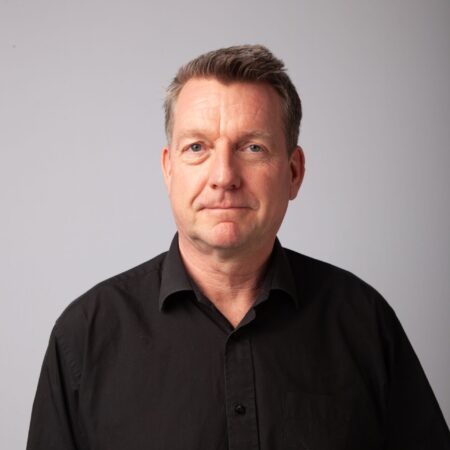 A middle-aged man with short brown hair, wearing a black button-up shirt, stands against a plain light grey background, looking directly at the camera with a neutral expression.