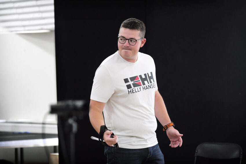 A man wearing glasses and a white Helly Hansen T-shirt stands indoors, holding something in his right hand, looking to his left. There is a black wall and some chairs in the background.