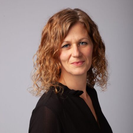 A woman with curly blonde hair, wearing a black blouse, stands in front of a plain light grey background and smiles slightly at the camera.
