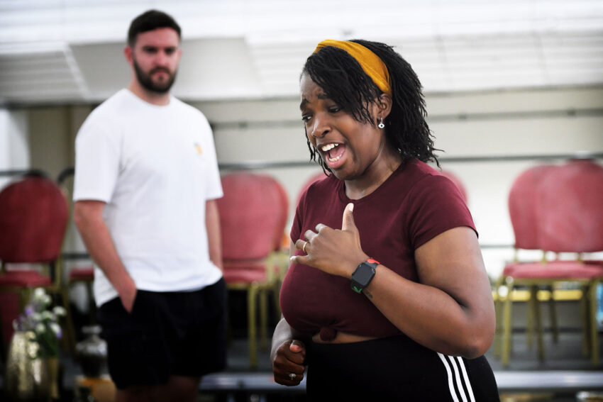 A woman in a maroon shirt passionately sings with one hand on her chest, whilst a man in a white shirt stands in the background near stacked red chairs.
