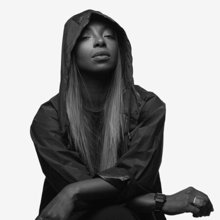 Afrodeutsche, an electronic music DJ, poses against a white background wearing a dark hooded jacket.