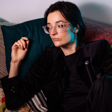 Anna Appleby with dark hair and glasses sits on a sofa, looking off to the side.