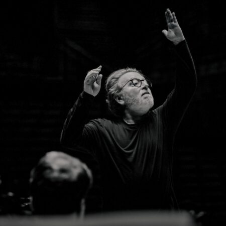 A conductor with grey hair and spectacles directing an orchestra with both arms raised.