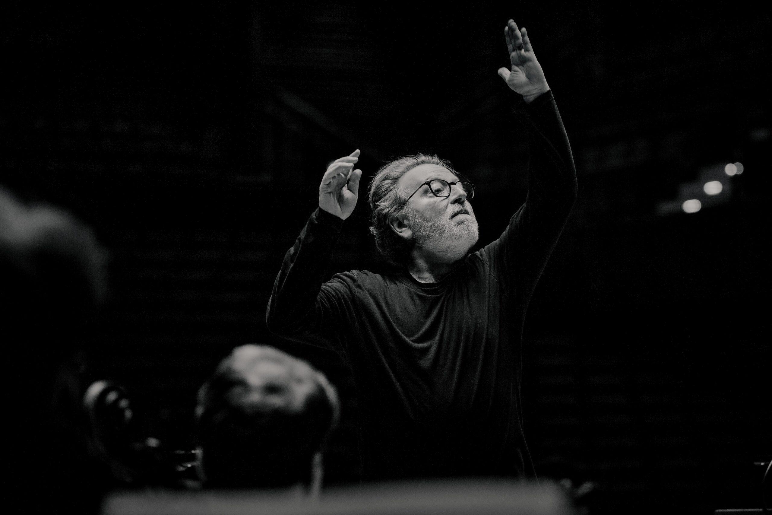A conductor with grey hair and spectacles directing an orchestra with both arms raised.