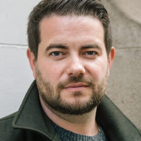 Headshot of a man with short dark hair and a beard, wearing a dark green collared coat over a grey jumper.