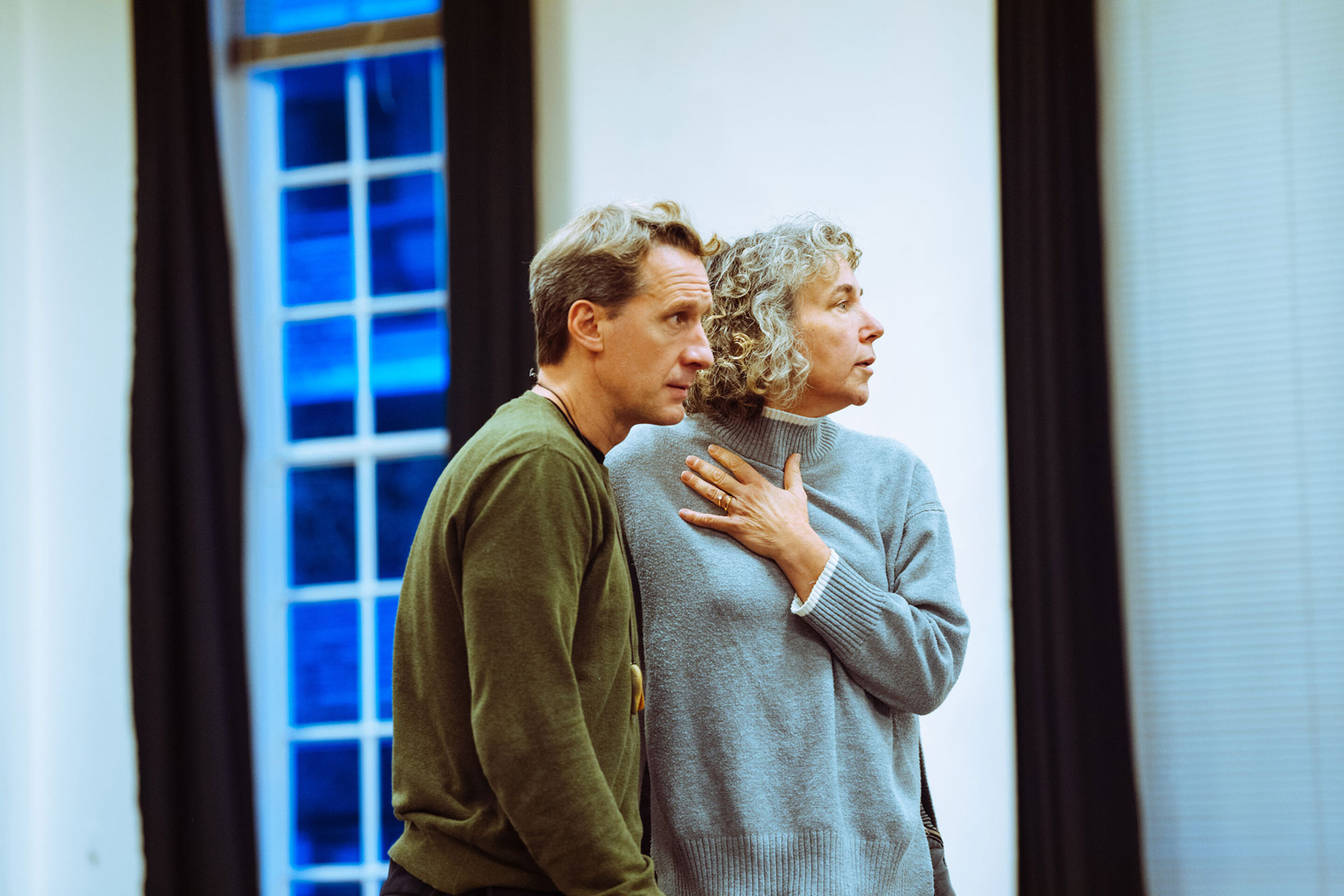 A man in a green jumper and a woman in a grey jumper stand side-by-side, both looking towards the right.