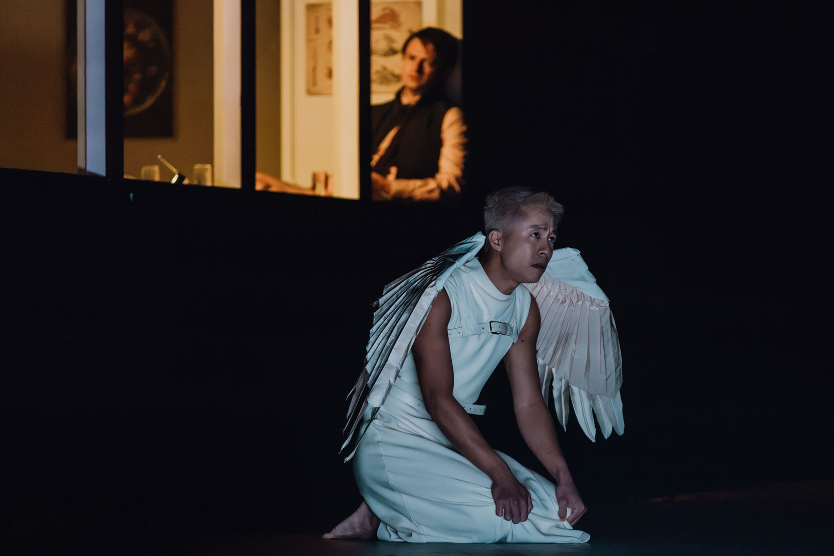 A person wearing white angel wings and a white sleeveless outfit kneels on a dark stage, whilst another person watches from a dimly lit doorway in the background.
