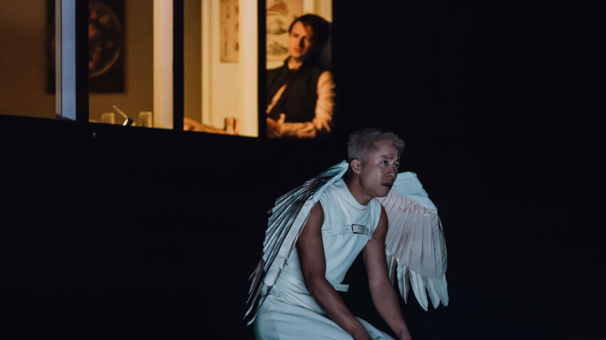 A person wearing white angel wings and a white sleeveless outfit kneels on a dark stage, whilst another person watches from a dimly lit doorway in the background.