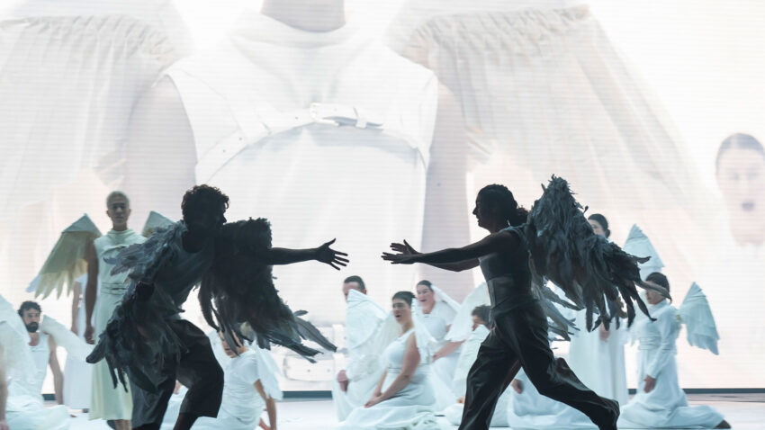 Two performers wearing feathered wings reach towards each other during a stage performance, surrounded by other costumed figures in front of a large projected image.
