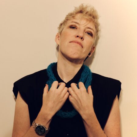A person with short blonde hair wears a black collared shirt and a chunky teal scarf.