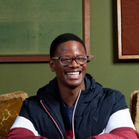 A man wearing glasses and a colour-blocked jacket laughing whilst sitting on a textured sofa.