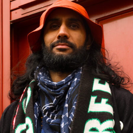 A person with a long beard and dark hair wearing an orange bucket hat, a blue patterned scarf, and a black and white scarf against a red background.