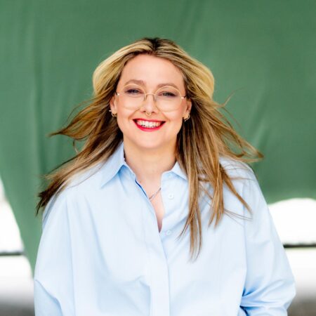 A smiling woman with blonde hair and round glasses wearing a light blue button-up shirt.