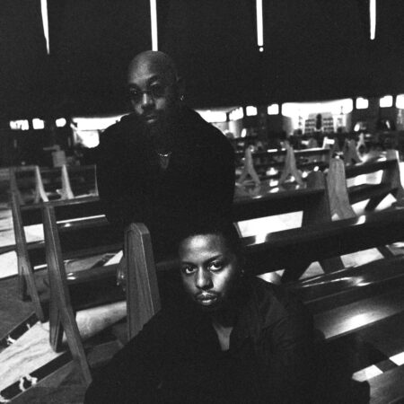 Two men in dark clothing pose together in a church among rows of wooden pews, capturing the ambient music aesthetic of Space Afrika.