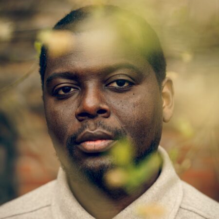 Close-up portrait of Richard Kudekha-Lark looking directly at the camera through out-of-focus foliage.